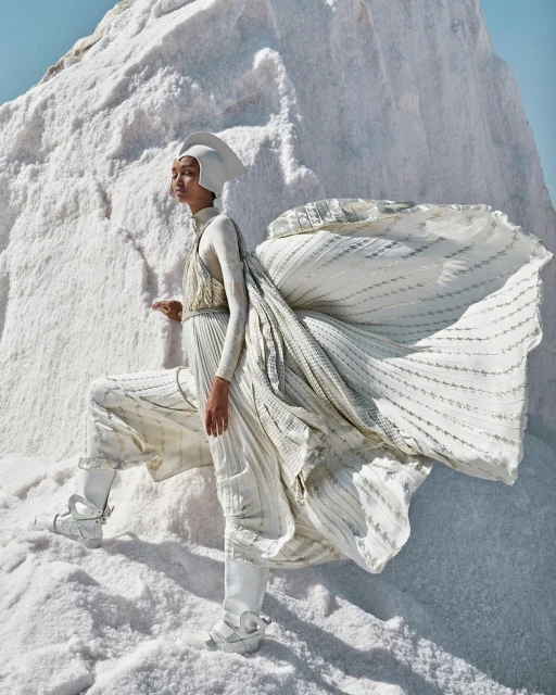 Fashion editorial shot of a woman in a white couture outfit and headpiece posing on a stark white textured salt flat landscape.