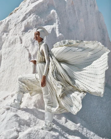 Fashion editorial shot of a woman in a white couture outfit and headpiece posing on a stark white textured salt flat landscape.