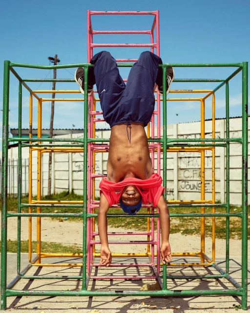 Young Black man hanging upside down on a colorful metal jungle gym. He is smiling with blue sky, wall, and a grassy outdoor setting.
