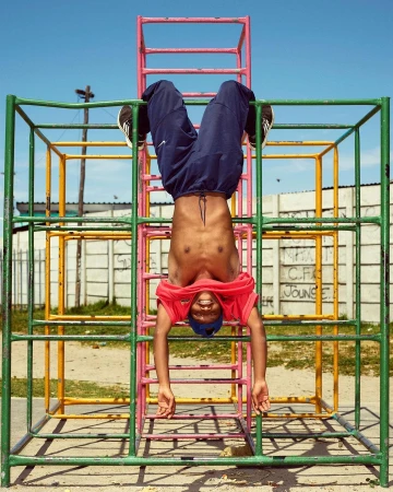 Young Black man hanging upside down on a colorful metal jungle gym. He is smiling with blue sky, wall, and a grassy outdoor setting.