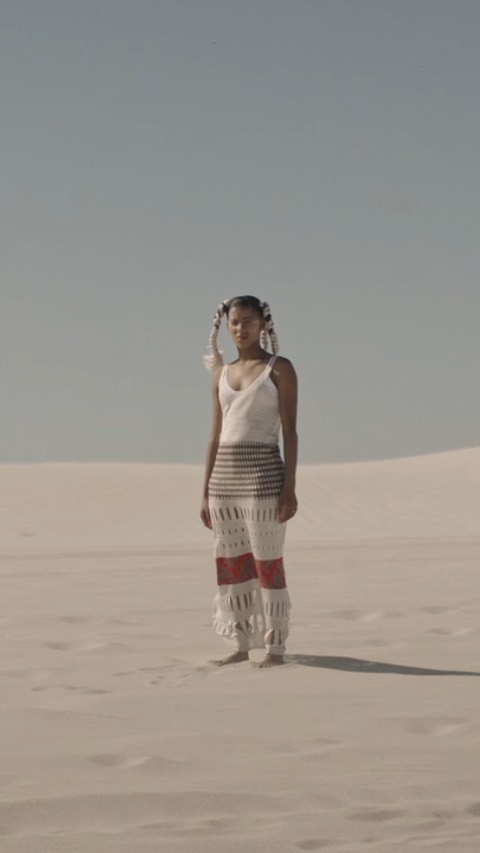 A woman stands in a sand dune landscape, wearing a white top and crocheted pants, with feathered hair accessories, under a clear sky.