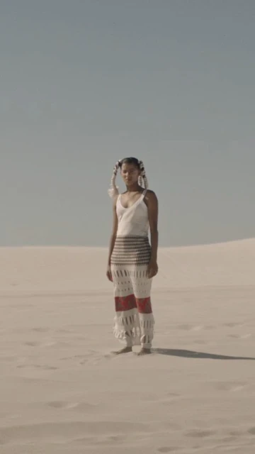 A woman stands in a sand dune landscape, wearing a white top and crocheted pants, with feathered hair accessories, under a clear sky.
