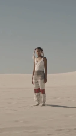 A woman stands in a sand dune landscape, wearing a white top and crocheted pants, with feathered hair accessories, under a clear sky.