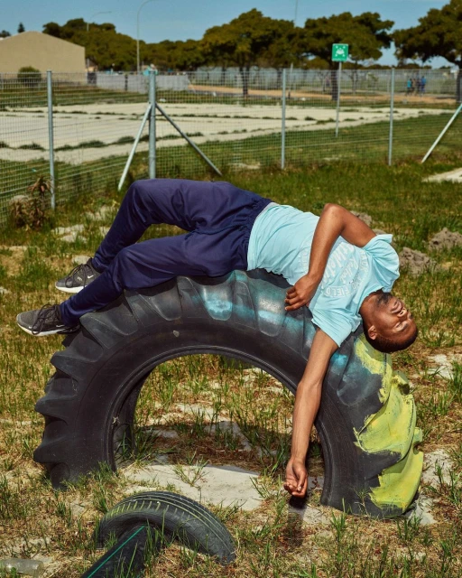 Young man in athletic wear rests on a tractor tire in a grassy, outdoor field with a distant building and blue sky on a sunny day.