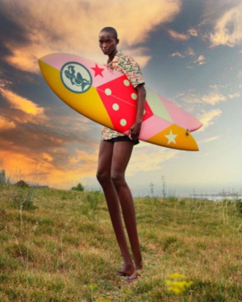 An adult woman with shaved head carries a colorful surfboard on a grassy hill under a dramatic sky, creating a striking outdoor scene.