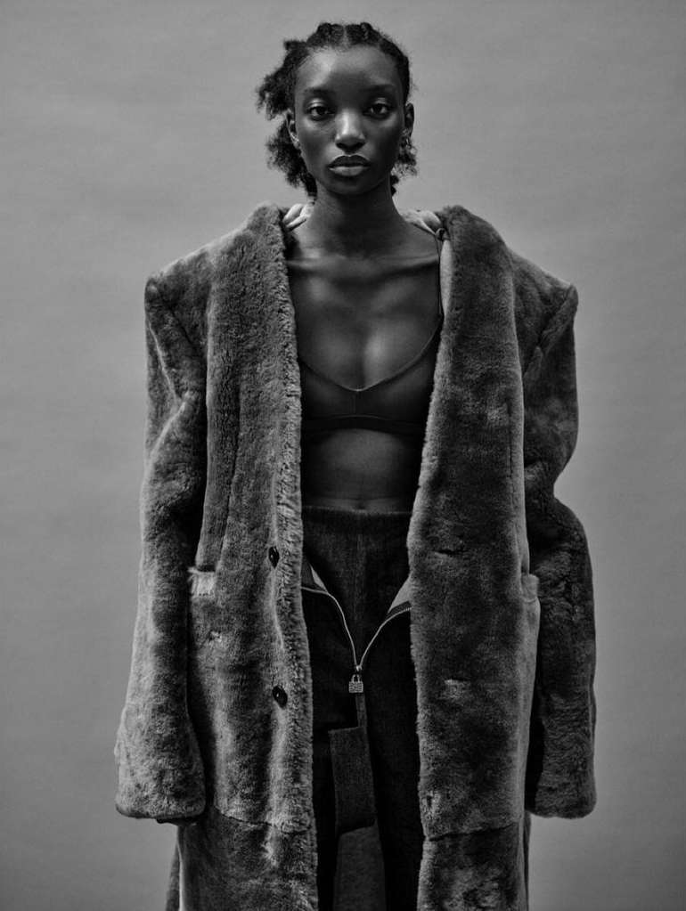 Black and white portrait of an adult woman in a fur coat, bra top, and trousers posing against a neutral gray background.