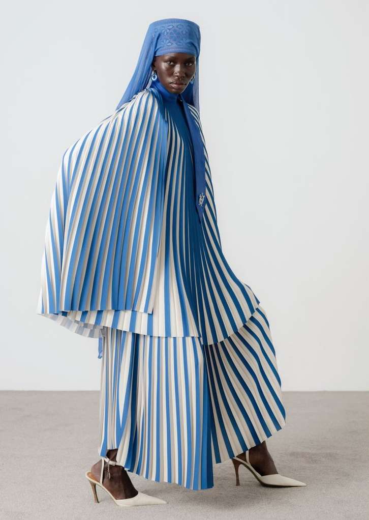 Fashion model in a blue and white pleated dress with head covering and earrings stands in a studio, wearing white high heels.