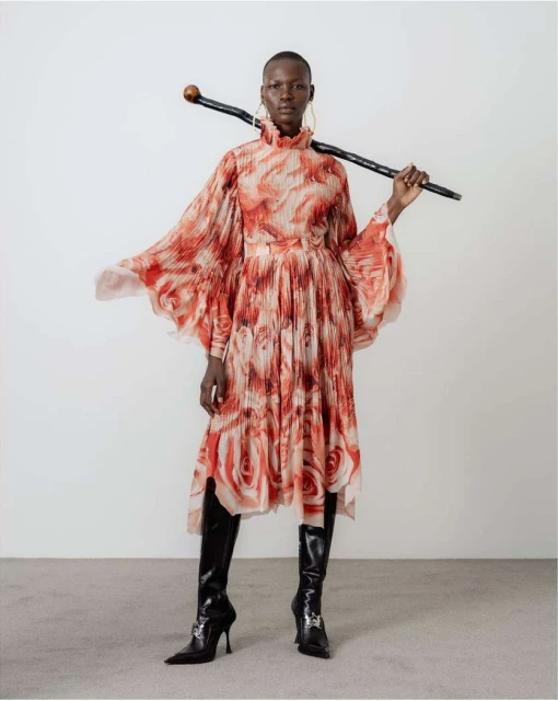 Elegant woman in a floral dress and boots poses with a walking stick against a white background in a fashion forward shot.