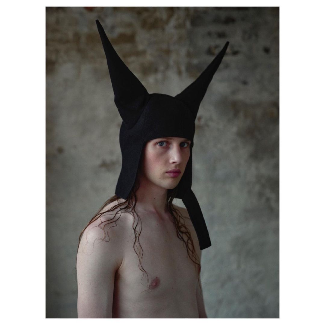 Androgynous person with bare chest and long hair wearing a pointy black headpiece with textural wall background in a medium studio shot.