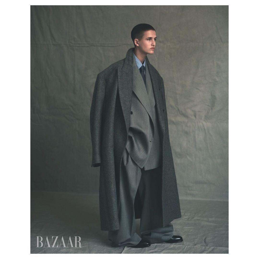 A person in a grey suit and oversized coat stands in front of a grey backdrop in a fashion editorial with the brand name Bazaar visible.