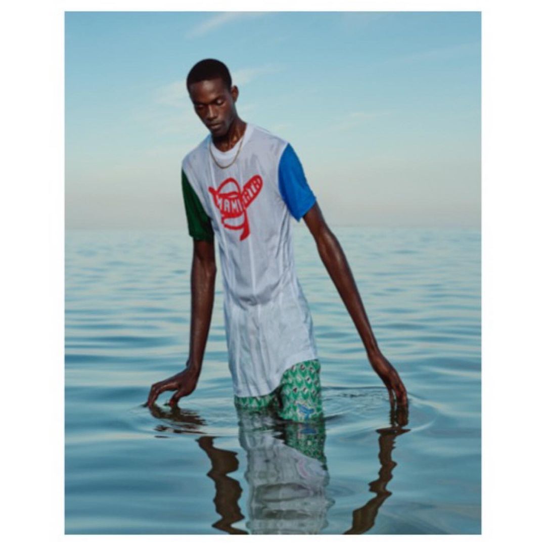 Dark-skinned man stands waist-deep in ocean wearing colorful shirt and swim trunks, with the horizon in the background on a clear day.