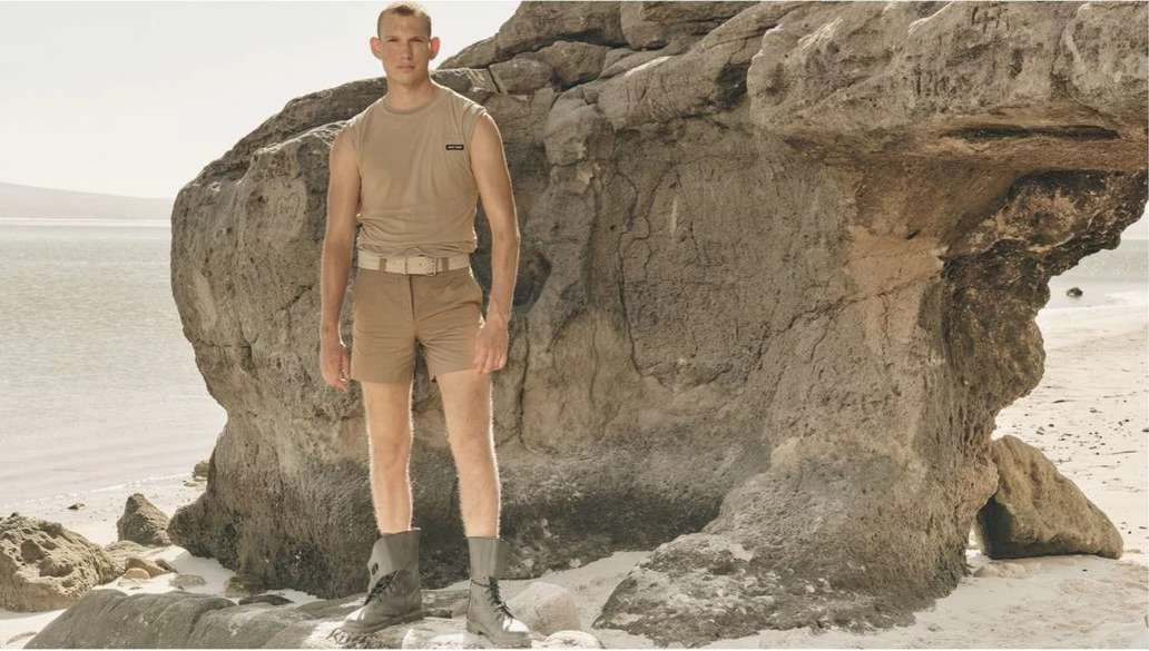 Male model wearing a beige top and shorts, posing next to a rock formation on a beach with water in the distance.