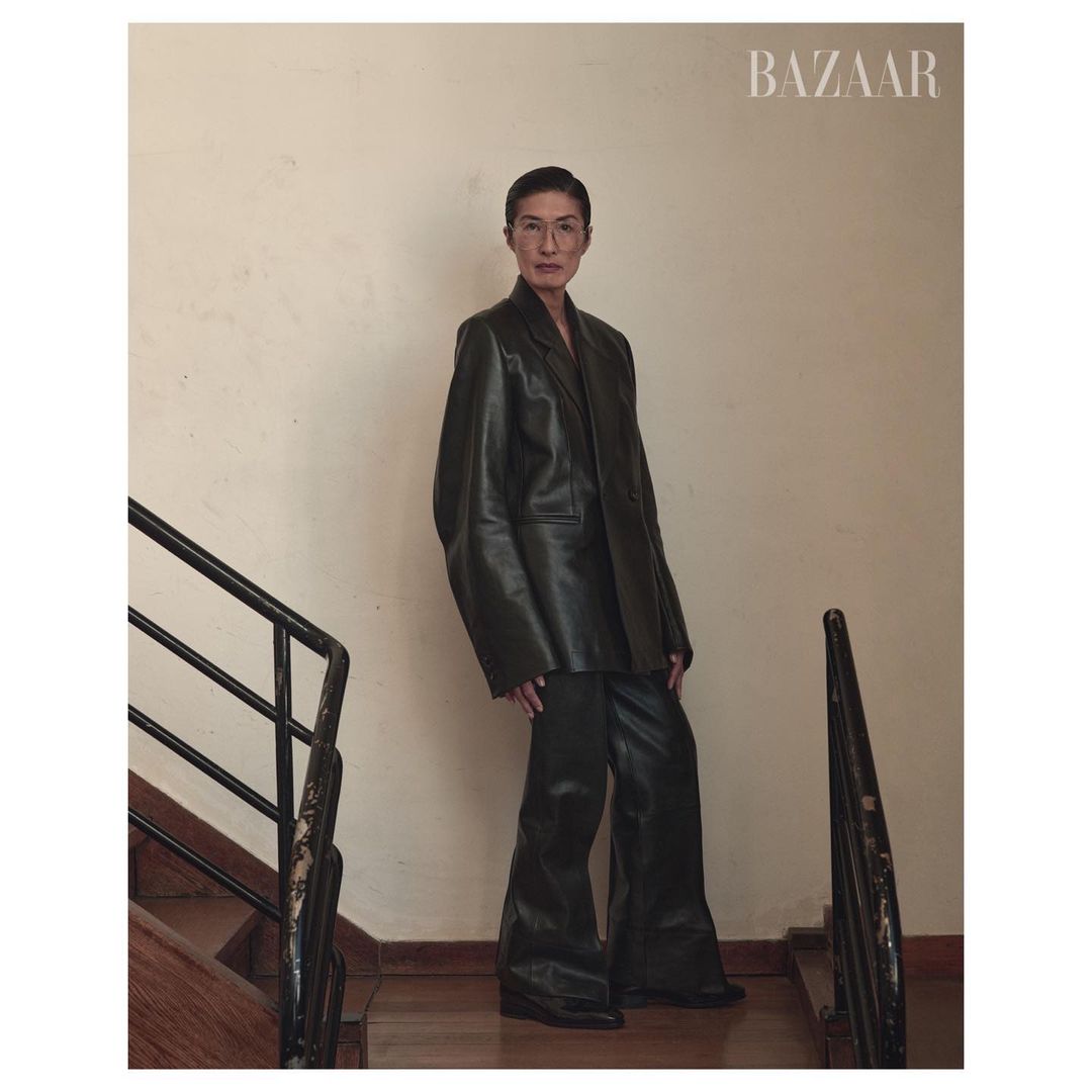 Fashion editorial shot of a stylish woman in an oversized black leather suit standing on wooden stairs in front of a beige wall.