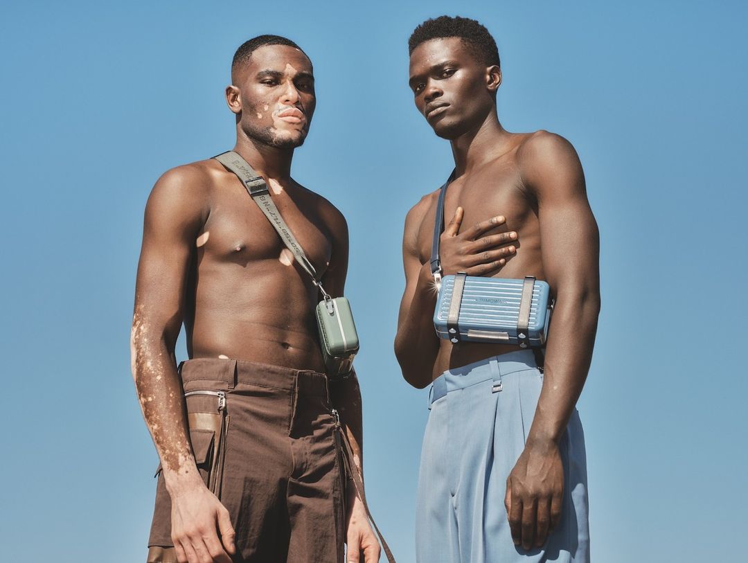 Two dark-skinned male models pose outdoors, wearing casual pants and crossbody bags against a clear blue sky in a fashion shoot.