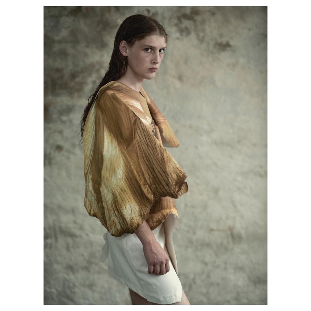 Young adult poses in a neutral studio backdrop. She's wearing a textured tan tunic and a white skirt. Wet hair adds to the dramatic presentation.