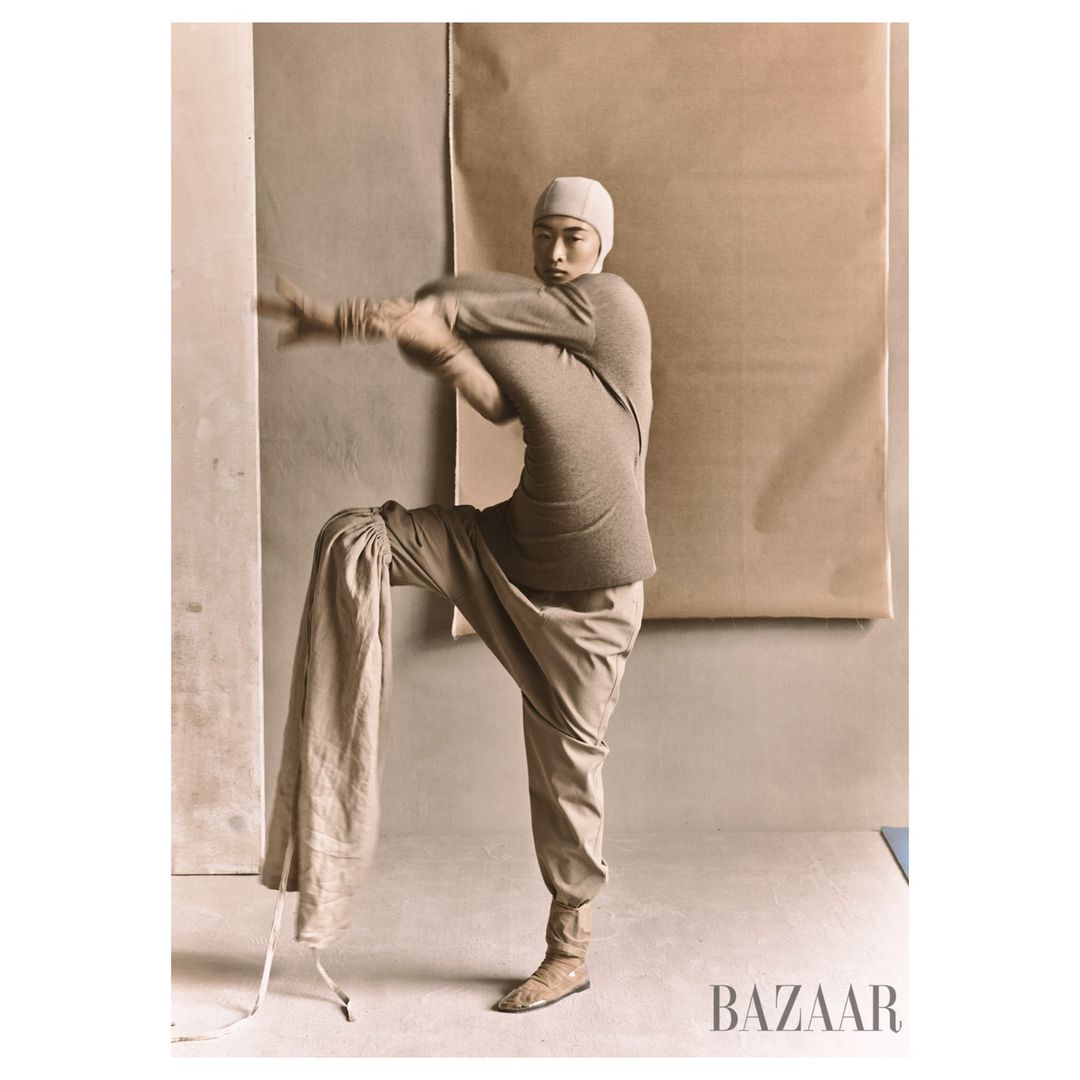 Fashion editorial shot of a person in monochrome outfit stretching with a beige backdrop and visible 'BAZAAR' logo in the corner.