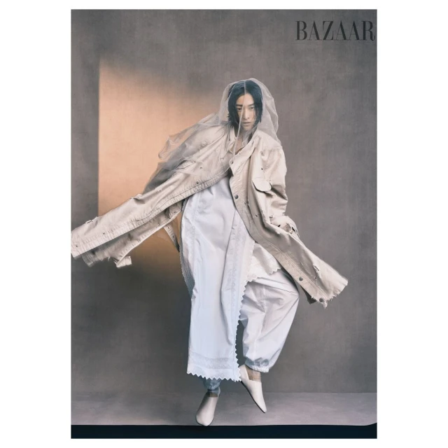 Fashion editorial shot of an Asian woman in white clothing and a tan overcoat, posing in front of a neutral background for Bazaar magazine.