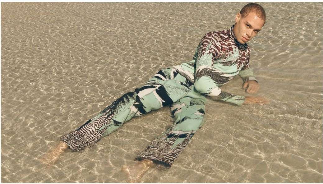 Adult male lying in shallow water wearing a patterned long-sleeve shirt and pants, looking directly at the camera with a neutral expression.