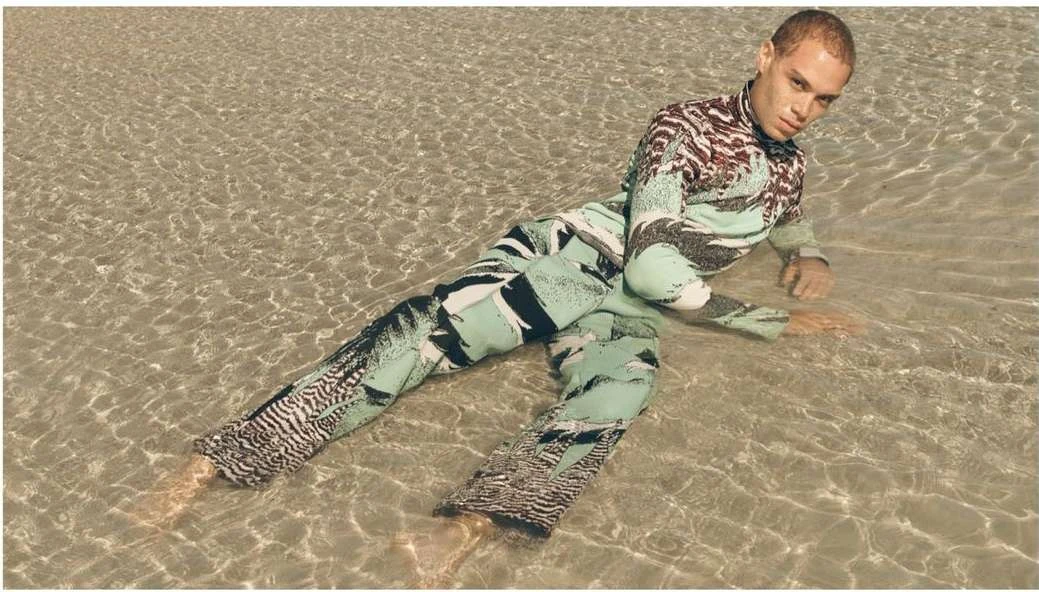 Adult male lying in shallow water wearing a patterned long-sleeve shirt and pants, looking directly at the camera with a neutral expression.