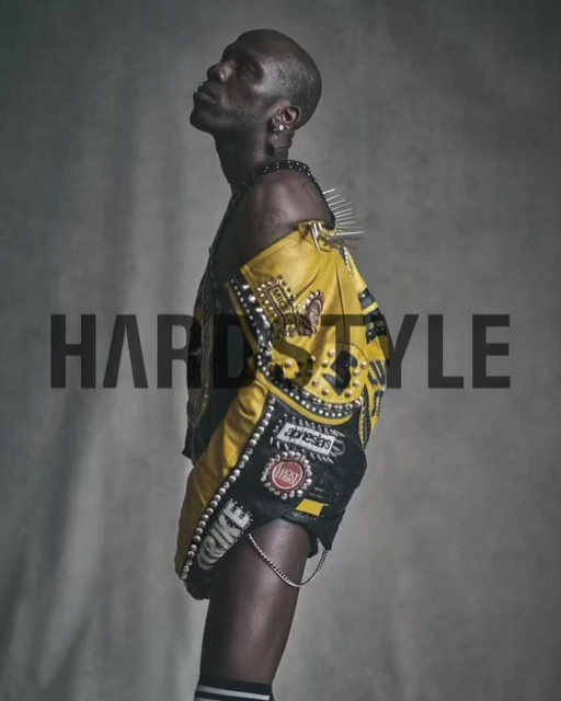 Stylish bald Black man poses in a yellow studded jacket and athletic shorts against a grey backdrop, showcasing an edgy, urban fashion look.