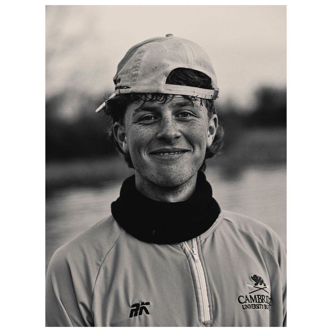 Portrait of a smiling young man wearing a baseball cap backward and a zipped jacket, captured in black and white.
