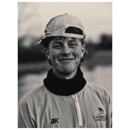 Portrait of a smiling young man wearing a baseball cap backward and a zipped jacket, captured in black and white.