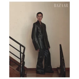 An older woman in a dark leather suit stands near a wooden staircase, photographed by Pieter Hugo in Cape Town.