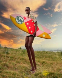 Man holding a brightly colored surfboard with star and diamond patterns against an outdoor sunset sky and grassy landscape