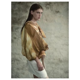 Portrait of a woman with dark hair wearing a brown patterned top and white skirt against a weathered concrete wall.