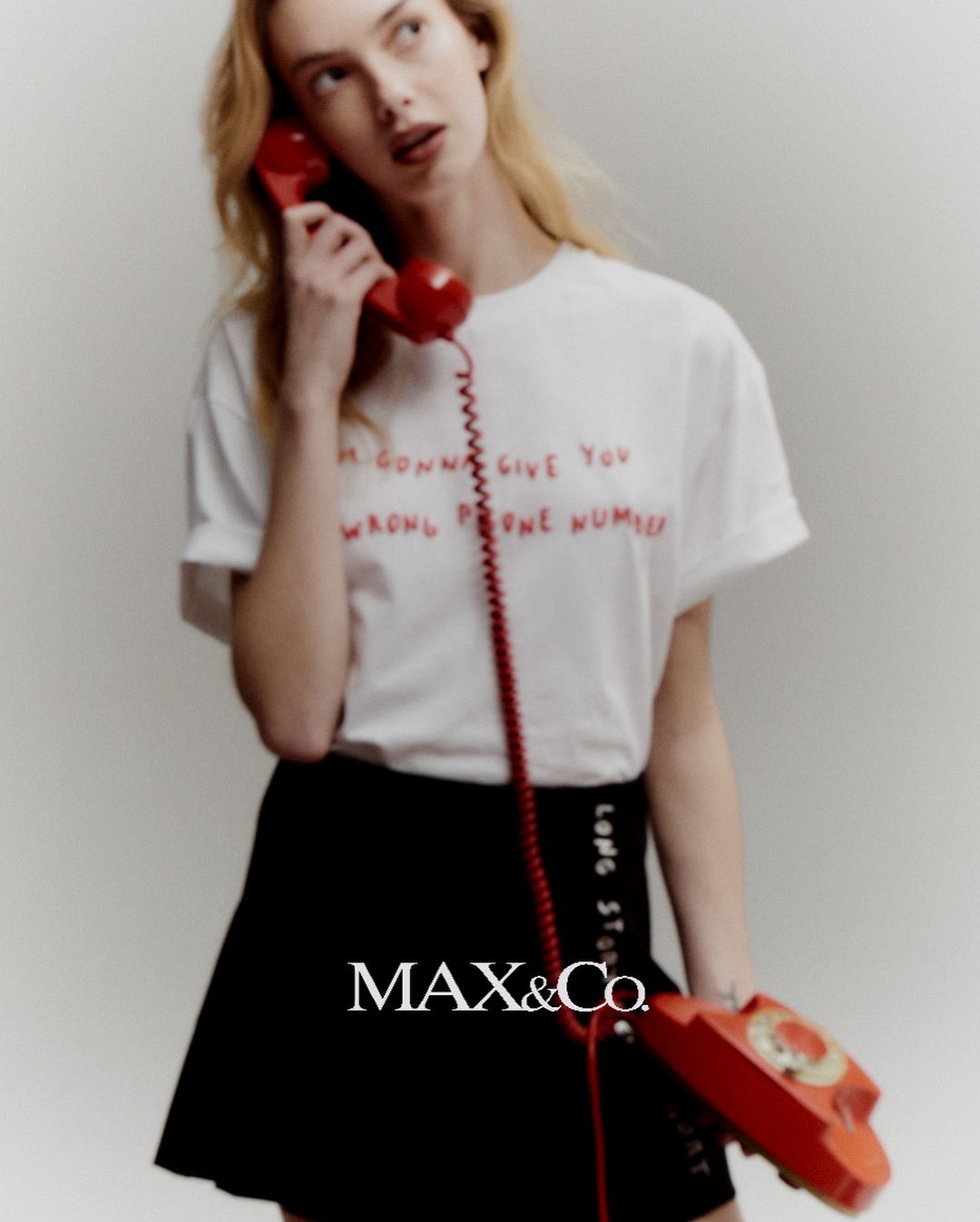 A young adult woman with light hair is on the phone wearing a t-shirt with red text, and a black skirt; she is posing in a studio.