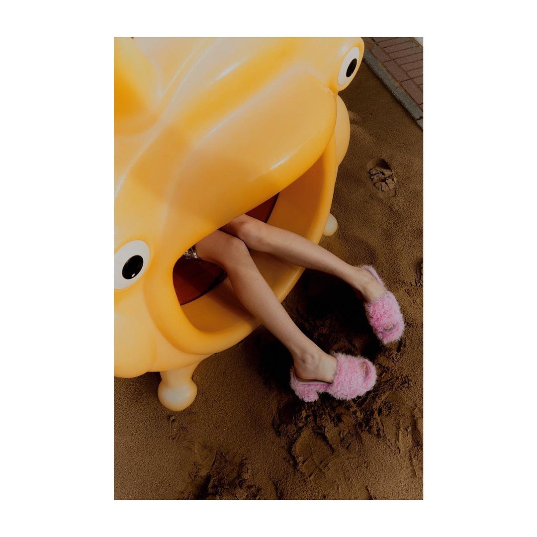 Child's legs and pink fuzzy slippers are visible inside an orange animal-shaped playground tunnel, with brown sand underneath.