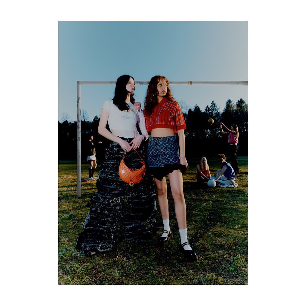 Two young women pose in sporty skirts and crop tops on a grassy field in front of a goalpost, with a group of people in background.
