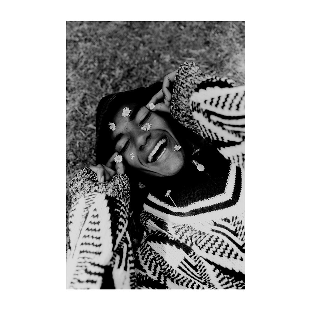 Smiling person with daisies on face lies on grass, wearing a patterned sweater. Black and white portrait captures a joyful moment.