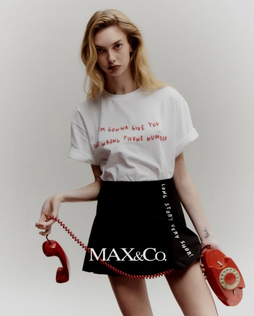 A fair-skinned woman with wavy blonde hair poses in a white t-shirt and black skirt, holding two red retro telephones in a light studio.