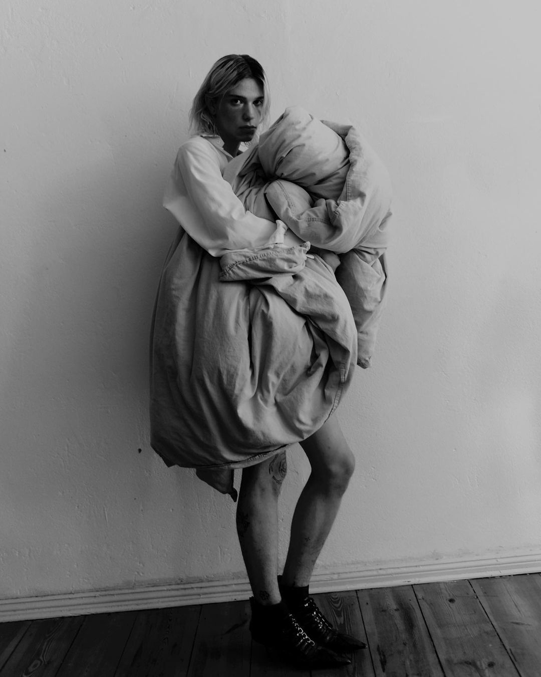 Monochrome shot of a blond adult woman holding a duvet, wearing a shirt and boots, posing against a plain wall on a wood floor.