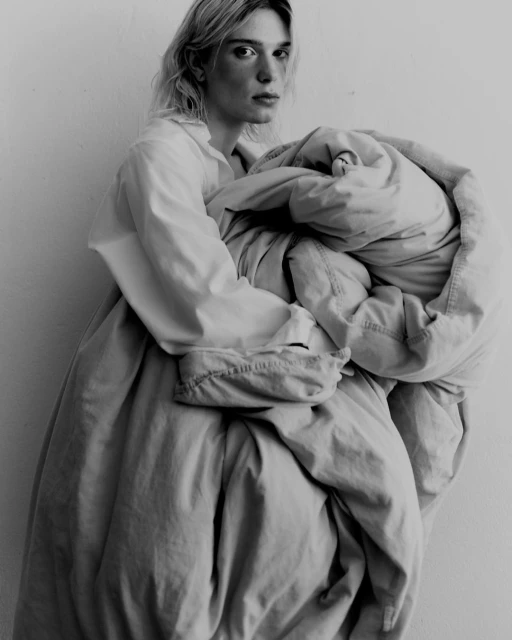 Black and white portrait of a fair-skinned adult with blonde hair, holding a large bundle of beige fabric indoors against a white wall.