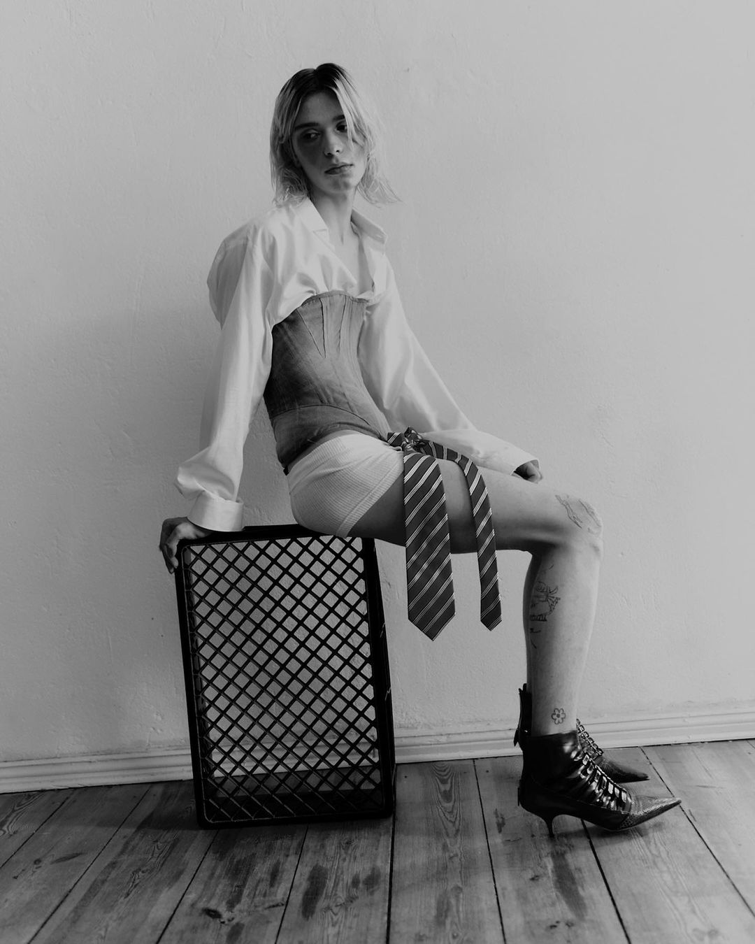 Black and white portrait of androgynous person sitting on a crate wearing a corset, collared shirt, boxer shorts, ties, and heeled boots.