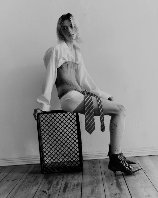 Black and white portrait of androgynous person sitting on a crate wearing a corset, collared shirt, boxer shorts, ties, and heeled boots.