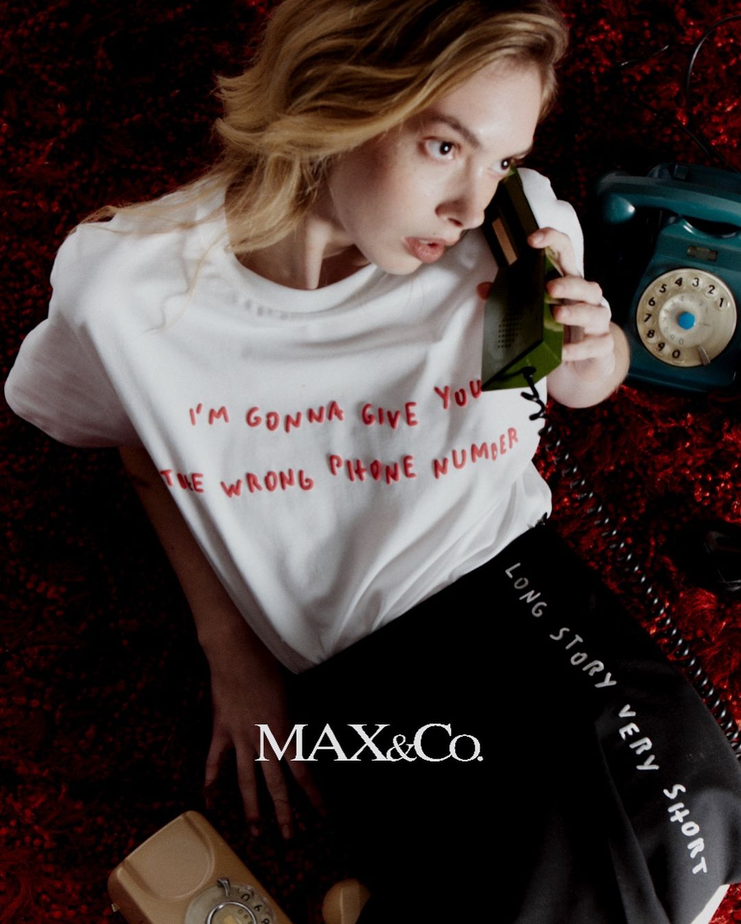 A blond woman wearing a white t-shirt and black skirt on a textured red carpet holding a green rotary phone.