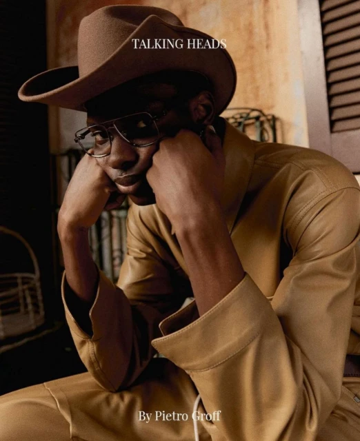 Adult male wearing a cowboy hat and glasses, posing in a tan outfit and looking at the camera in an indoor environment.