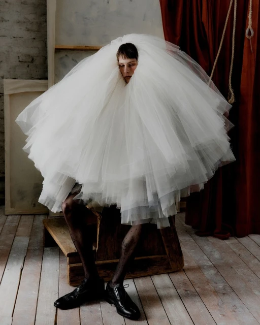 A person sits on a wooden block wearing black shoes and a large white tulle dress in a photo studio with soft lighting.