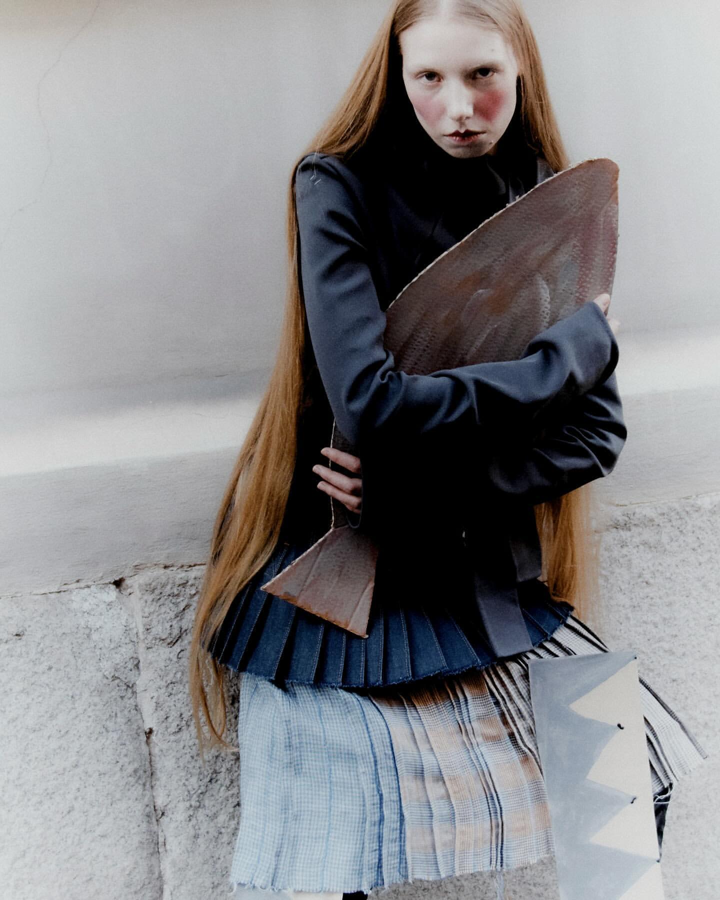 Adult woman with long red hair holds a sculptural object in a high-fashion editorial shot against a grey concrete wall backdrop.