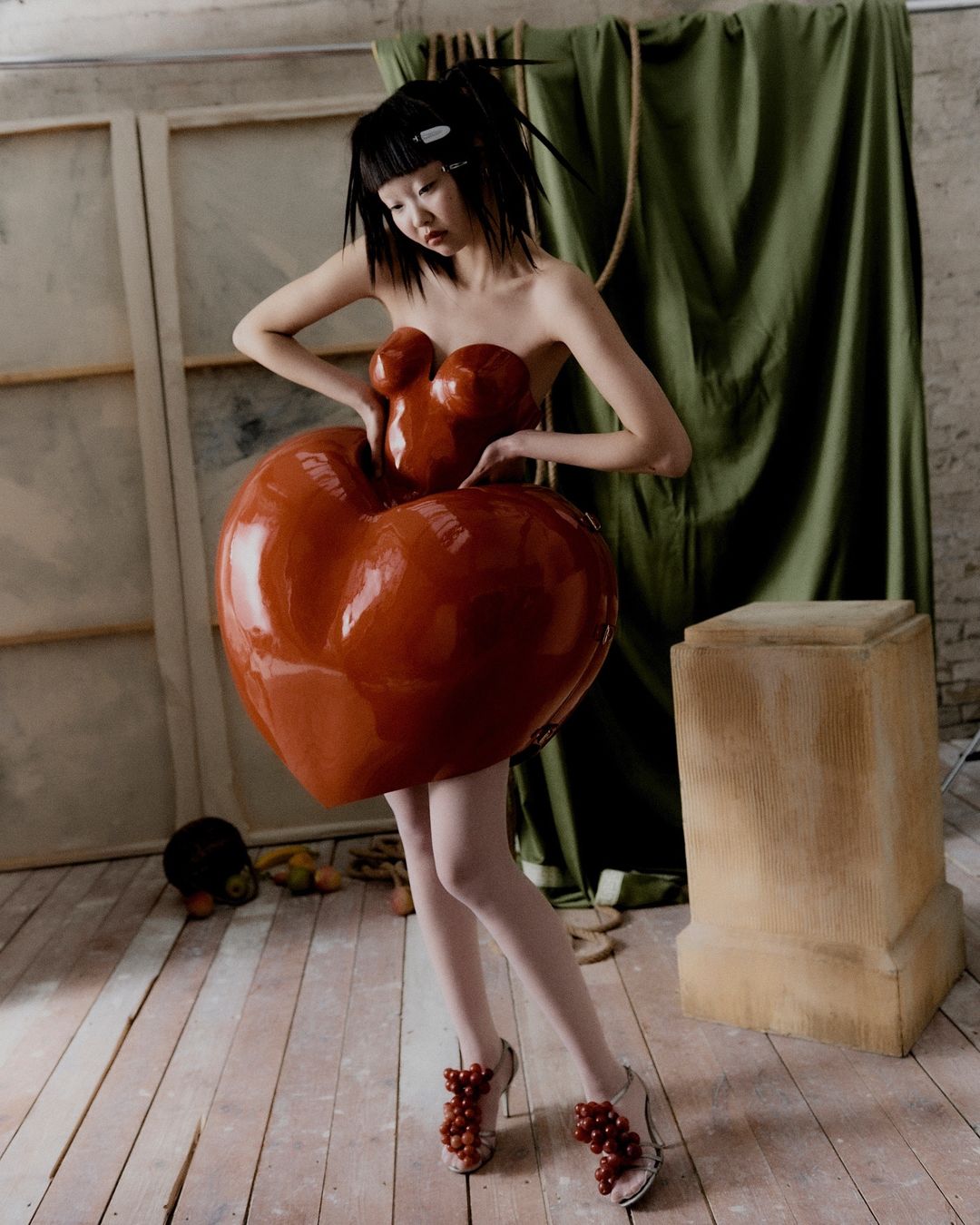 Asian woman poses in avant-garde red dress and fruit-covered sandals in a photo studio against a draped green fabric backdrop.