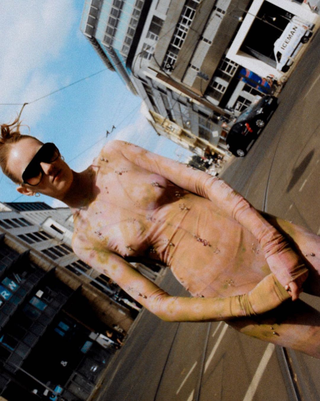 Fashionable woman in patterned bodysuit, sunglasses, and red hair poses on city street with buildings under blue sky on sunny day.