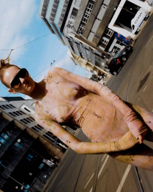 Fashionable woman in patterned bodysuit, sunglasses, and red hair poses on city street with buildings under blue sky on sunny day.