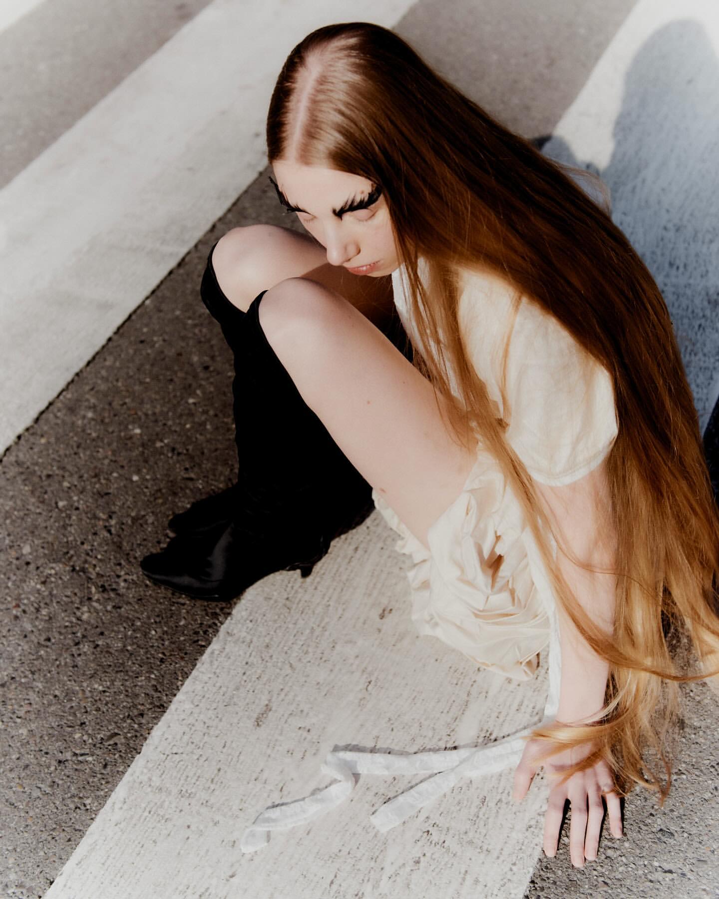 High angle view of a young woman with long red hair and dramatic false eyelashes posing while seated outdoors on concrete.