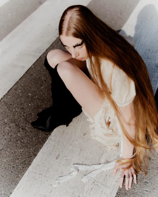 High angle view of a young woman with long red hair and dramatic false eyelashes posing while seated outdoors on concrete.