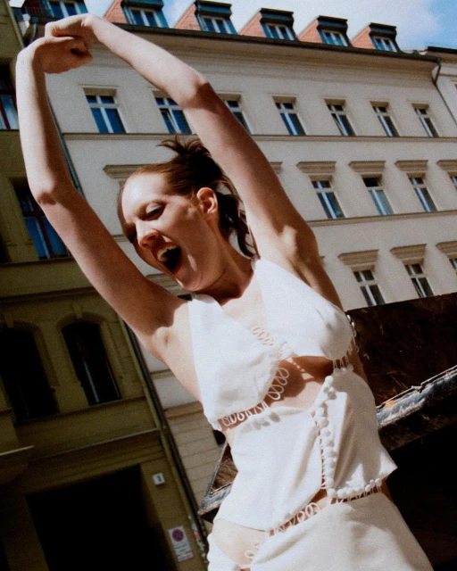 A stylish adult female with arms raised, standing outdoors in a custom white halter top and skirt with architectural background.