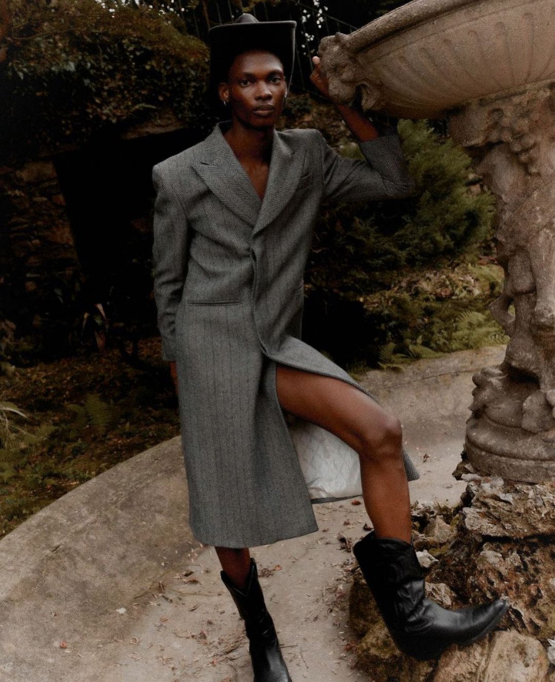 An androgynous person in a gray coat poses outdoors next to a stone sculpture wearing black boots and a hat in a natural outdoor setting.