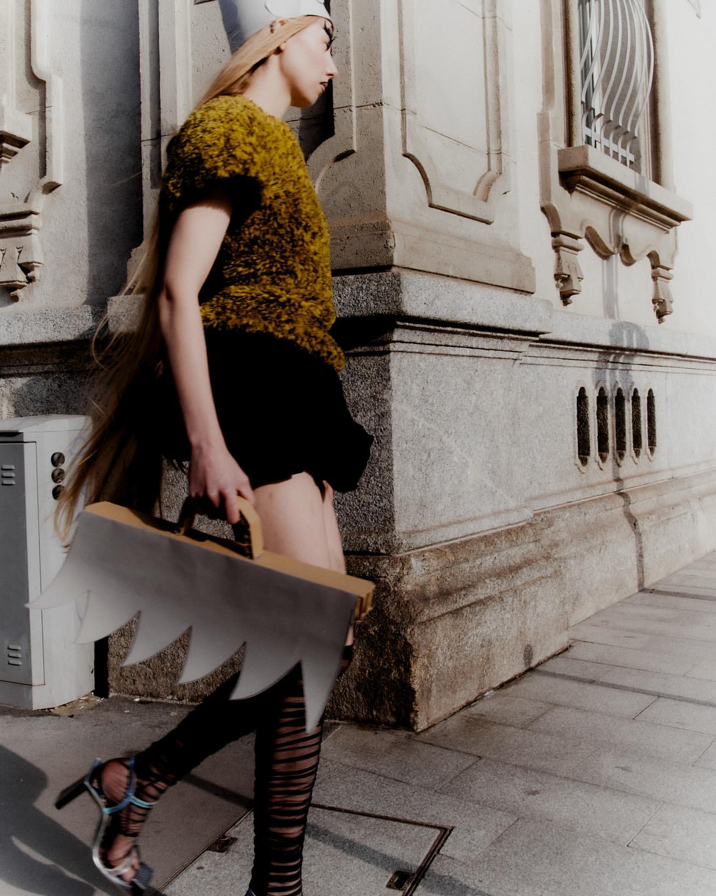 Fashionable adult female walks outdoors wearing a gold textured top, black skirt, patterned tights, and silver heels, carrying a unique prop.